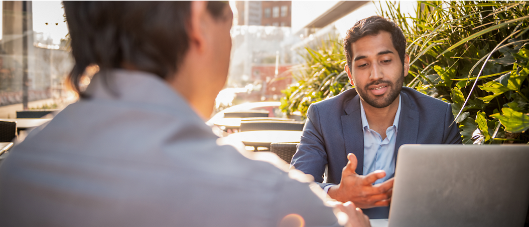 Two men engaging in a conversation at an outdoor café table, one gesturing while sitting behind a laptop.