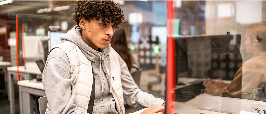 Un jeune homme aux cheveux frisés assis à un bureau d’ordinateur dans un bureau, détournant les yeux pensivement.