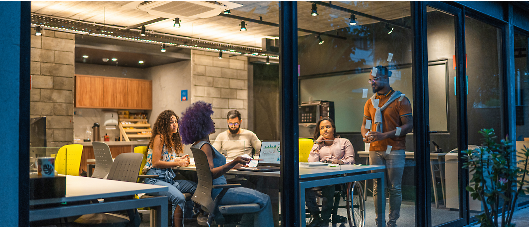 A diverse group of professionals in a meeting at a brightly-lit office space, with one person presenting to three others