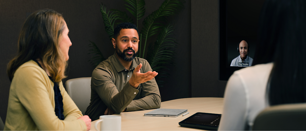 Two people in a meeting room discussing with a colleague who is participating via video call on a screen.