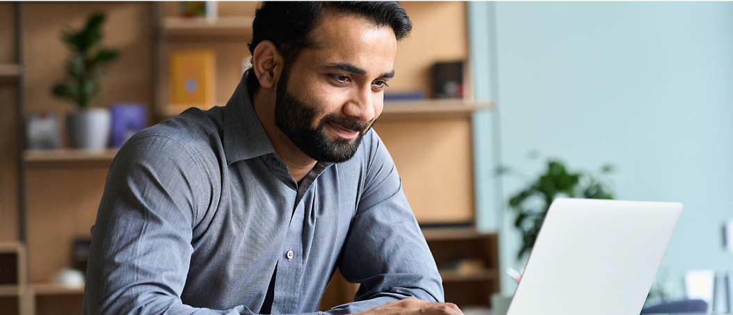 Un homme portant une barbe travaillant sur un ordinateur portable dans un environnement de bureau moderne, souriant devant l’écran.