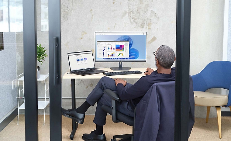 Una persona sentada en una oficina de cristal utilizando un portátil conectado a un monitor de escritorio