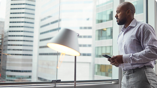 A man stands near a large window in an office holding a smartphone, looking outside at a cityscape with tall buildings.