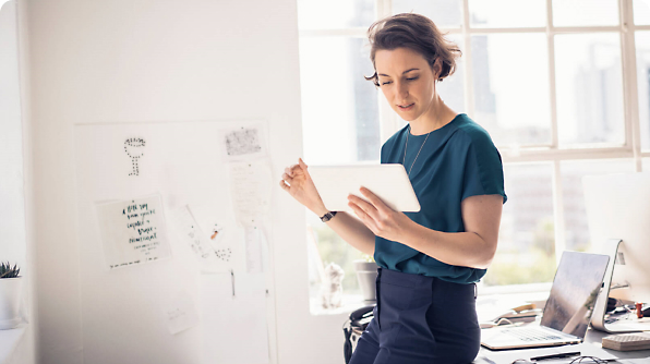 A woman stands in an office holding a tablet. Behind her is a whiteboard with papers.