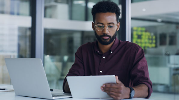 A man with glasses is sitting at a desk using a tablet with a laptop next to him.