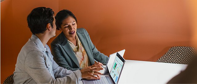 Two professionals discussing over a laptop in an office with orange walls.