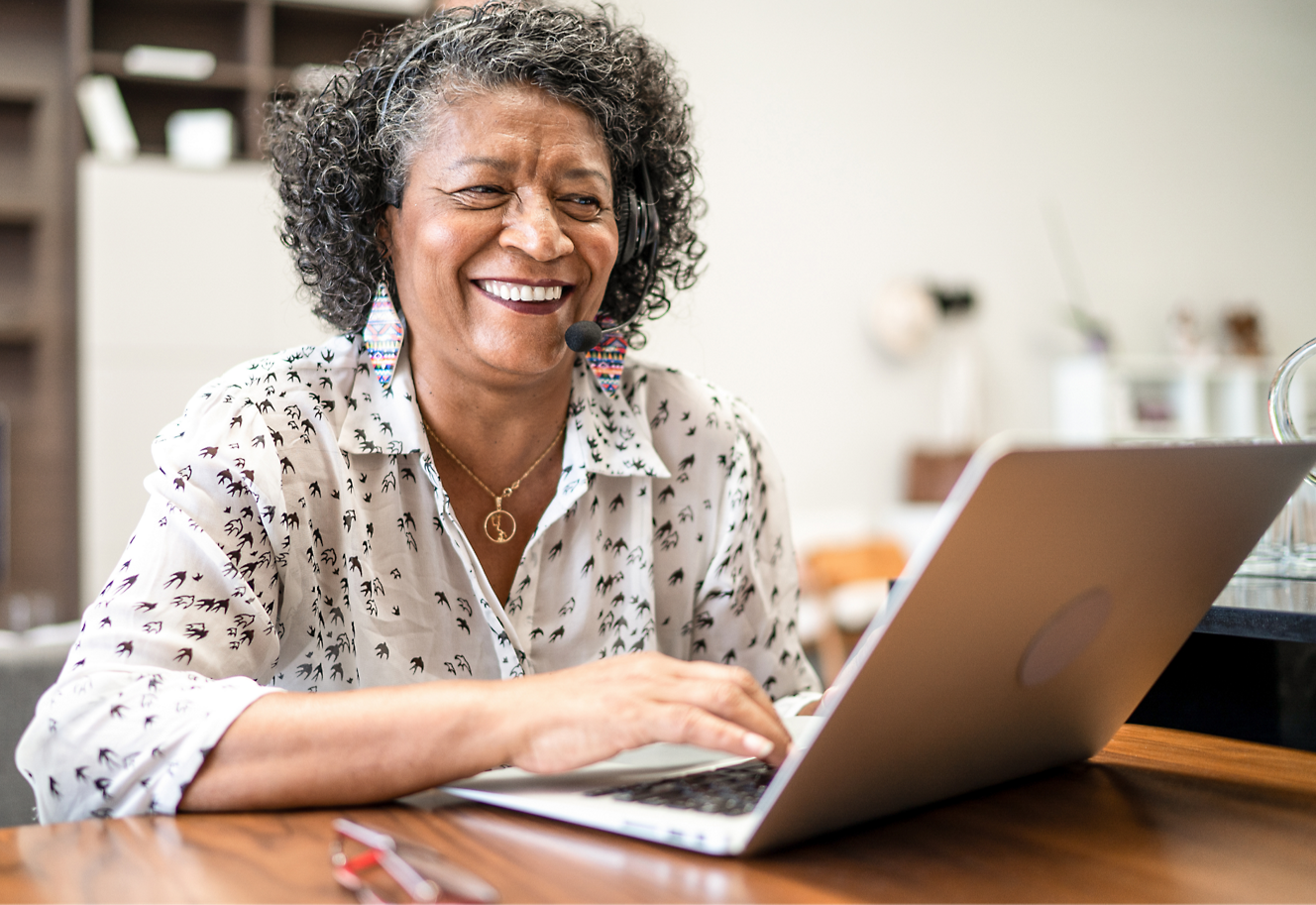 An elderly woman with curly hair is smiling while using a laptop.