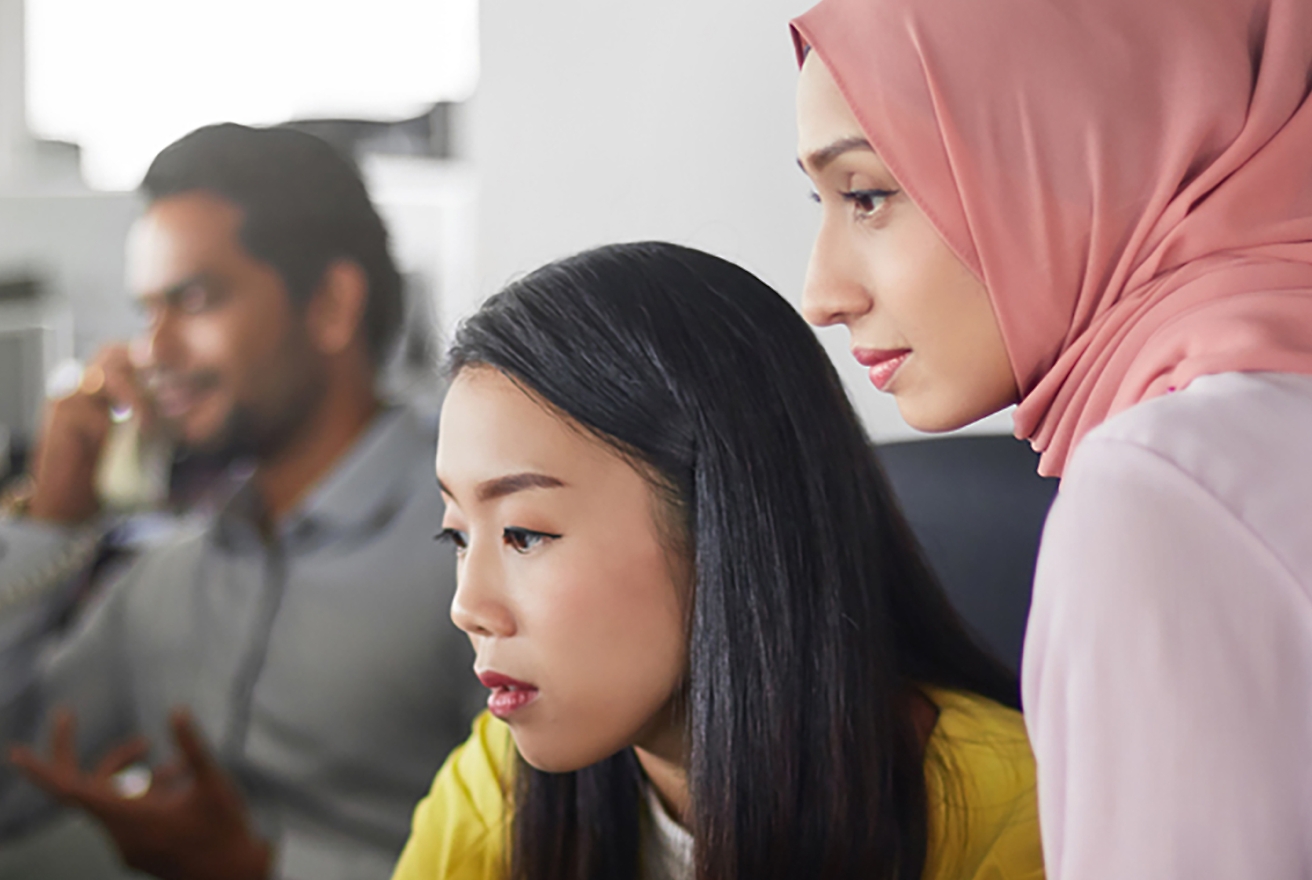 Two women, one in a hijab, closely observe a computer screen while a man speaks on a phone in the blurred background.