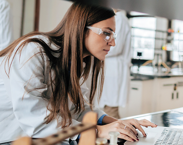 A woman wearing white lab coat and glasses is working on laptop