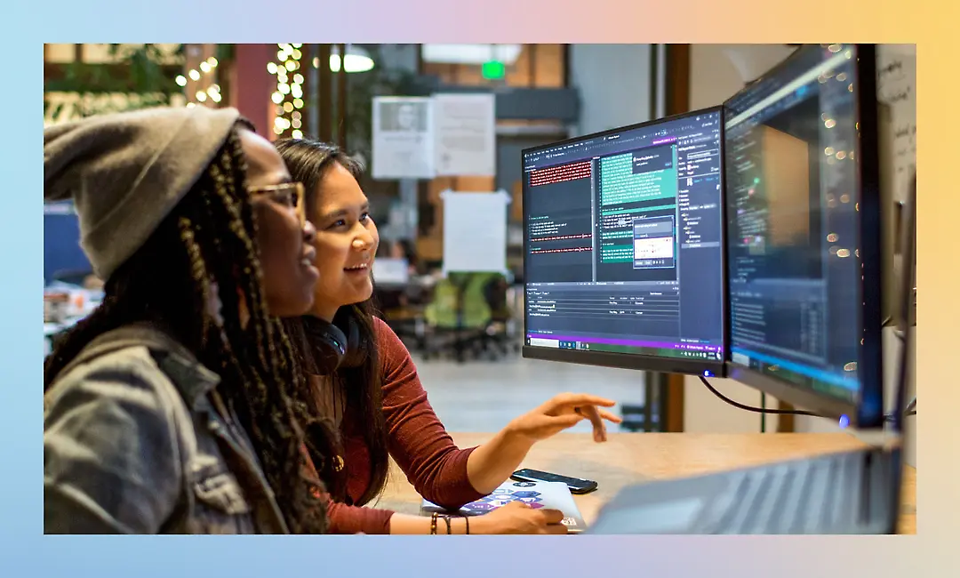 Two people are sitting at a desk with two large computer monitors, discussing the code displayed on the screens