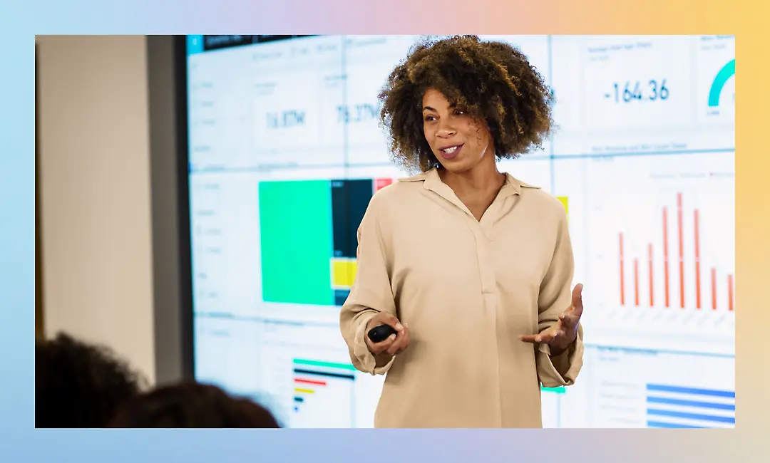 A person stands in front of a large screen displaying charts and graphs, holding a remote and speaking to an audience.