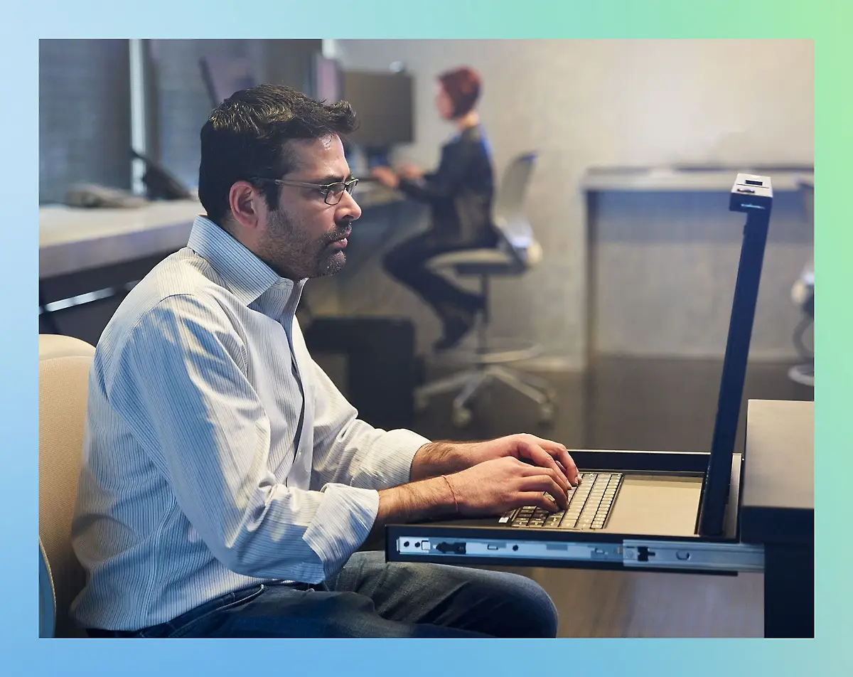 A man seated at a desk, focused on his laptop, surrounded by a tidy workspace and soft lighting.