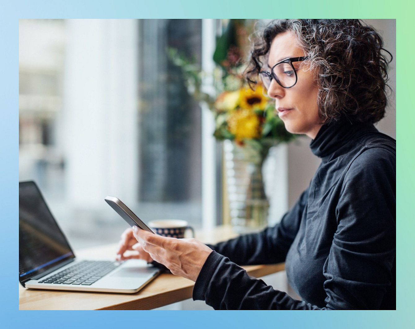 A person with curly hair and glasses sits at a table in front of a laptop, looking at a smartphone.