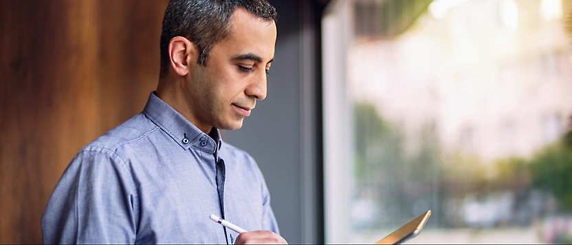 A person with short hair wearing a button-up shirt stands near a window, looking down at a tablet or notebook.