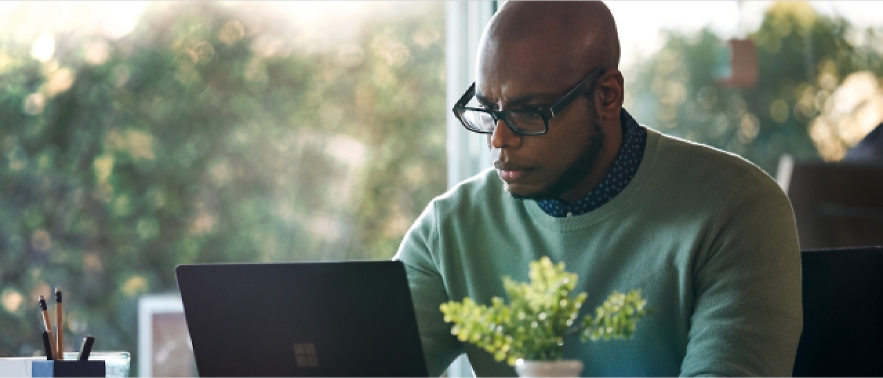 A person wearing glasses and a green sweater is focused on using a laptop at a desk with a small plant and a cup of pencils.