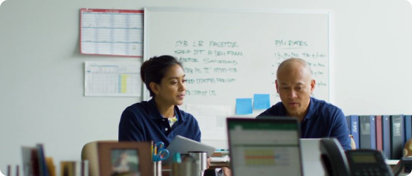 Two people sitting at a desk in front of a whiteboard.