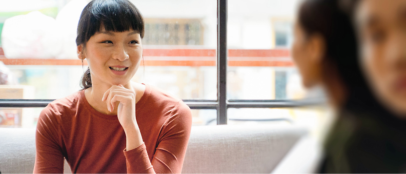 A person with dark hair and an orange top sits indoors, smiling and looking toward another person who is slightly blurred 