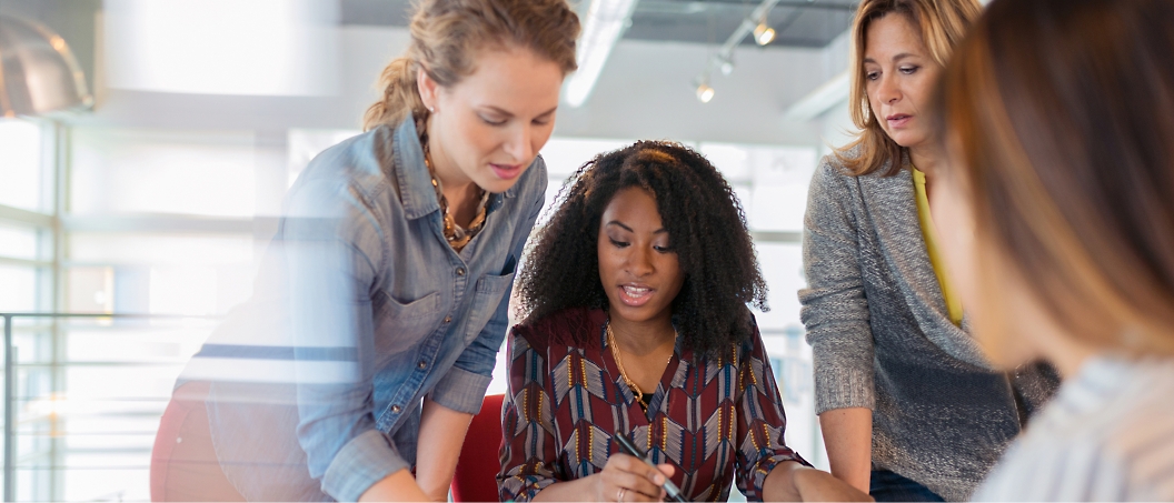 Four women collaborate at a table, reviewing documents and discussing work in a brightly lit office space.