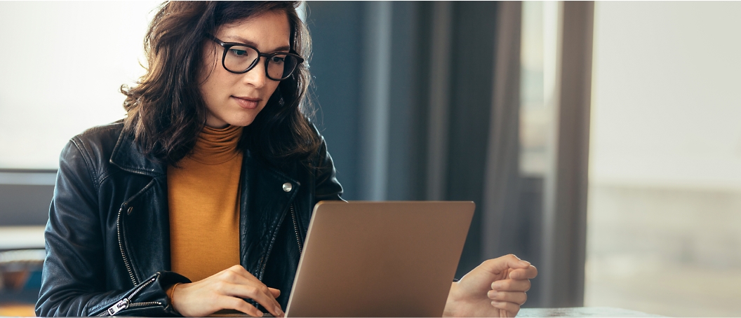 A person with glasses and dark hair is sitting at a table, focused on using a laptop.