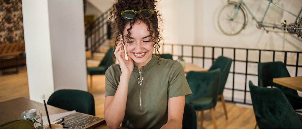 A person with curly hair in a green top smiles while talking on a phone in a modern café 