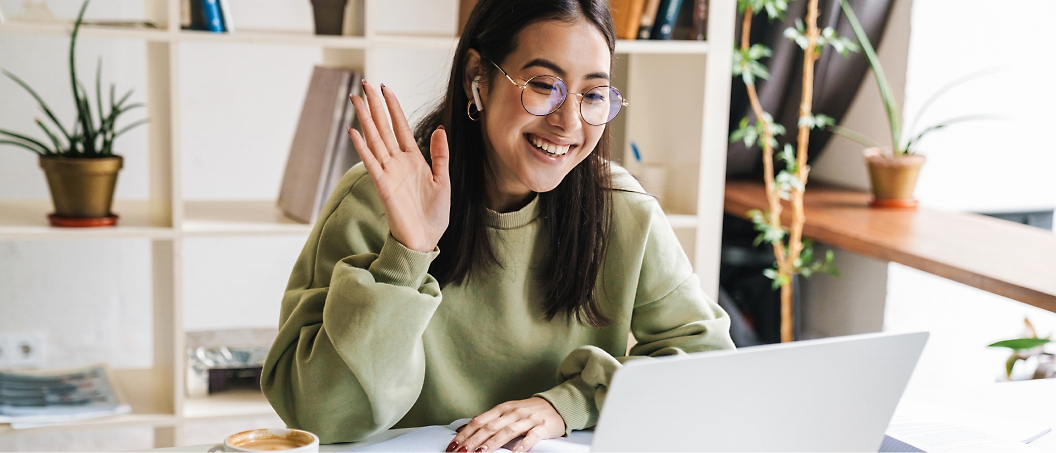 A woman wearing glasses and a green sweater waves at her laptop screen while sitting at a desk with a cup of coffee. 