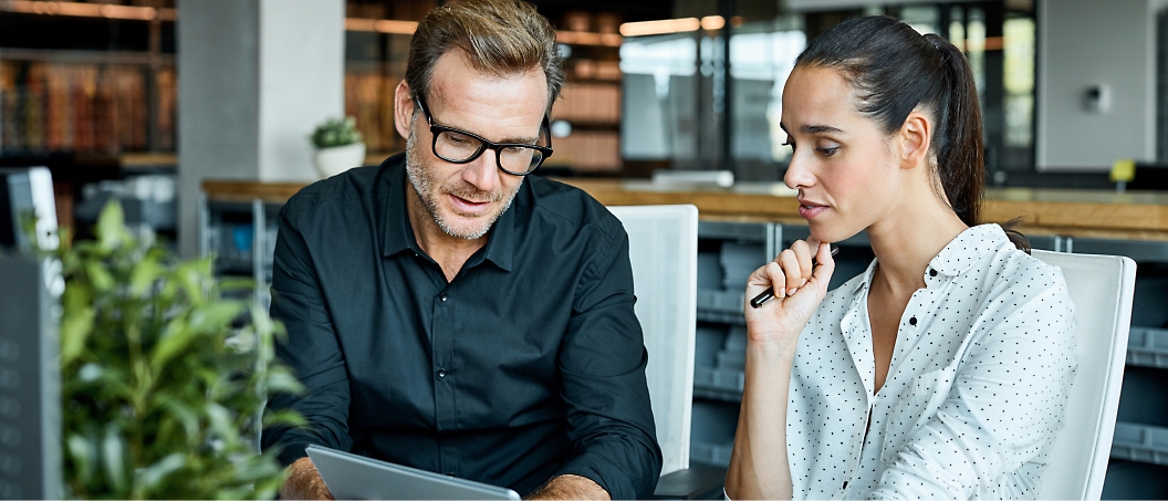 Two colleagues, a man and a woman, sit at a desk in an office and discuss something on a tablet. 