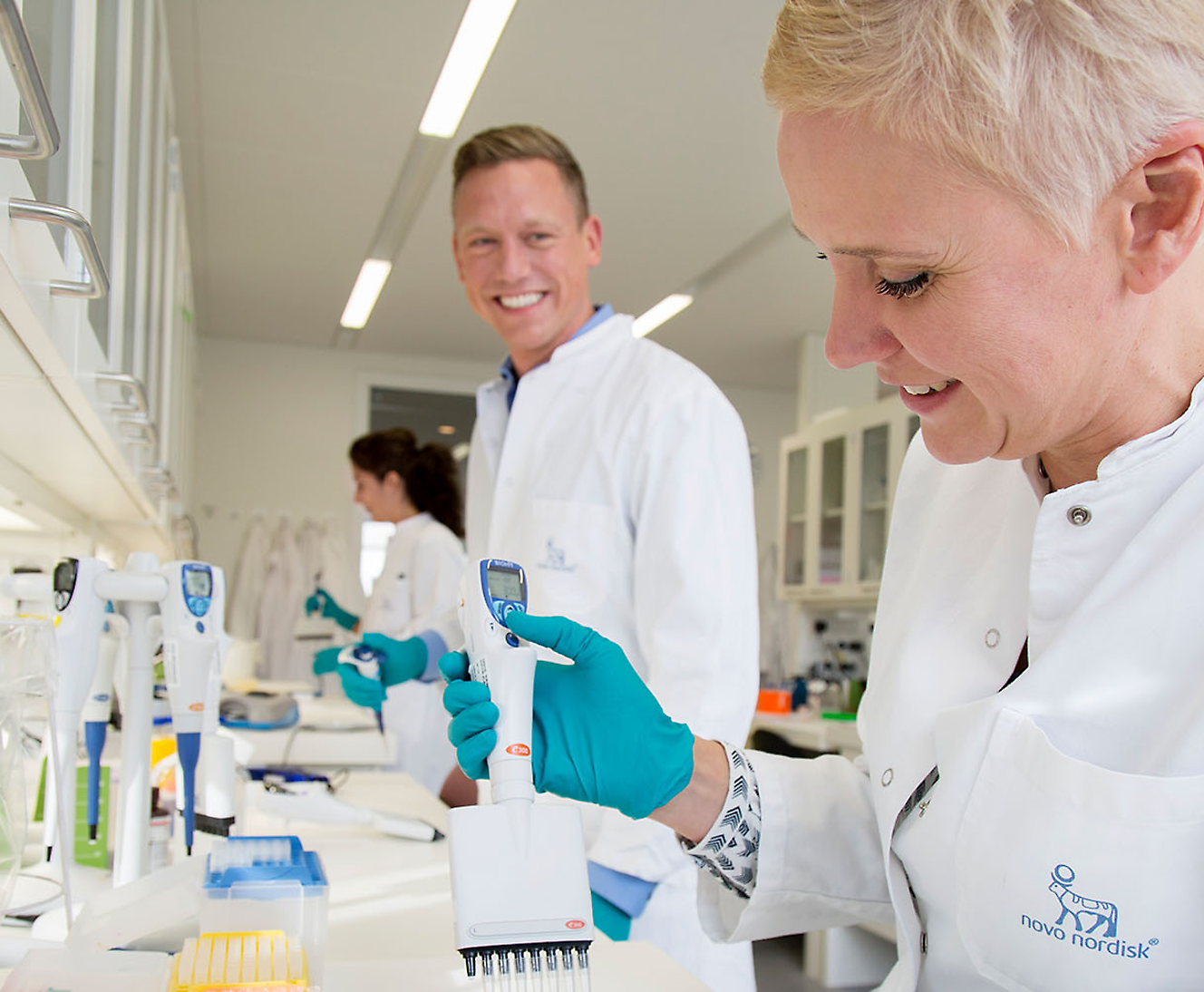 Three scientists wearing lab coats and gloves work in a laboratory. One woman in the foreground uses a pipette while smiling.