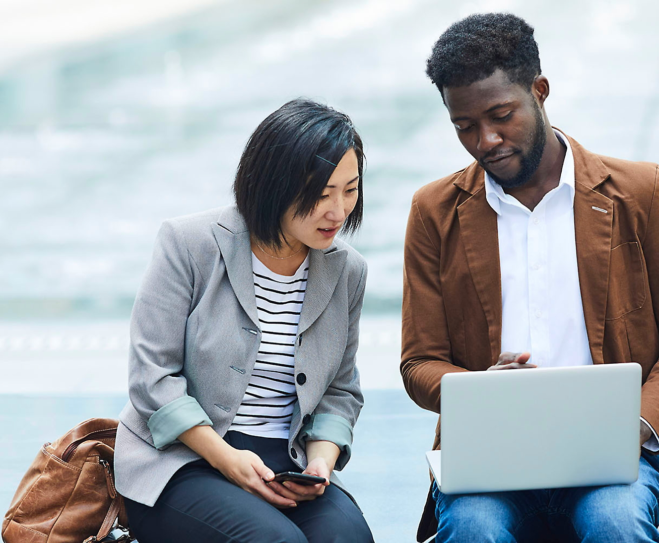 Two people sit outdoors with a laptop; one appears to be explaining something to the other