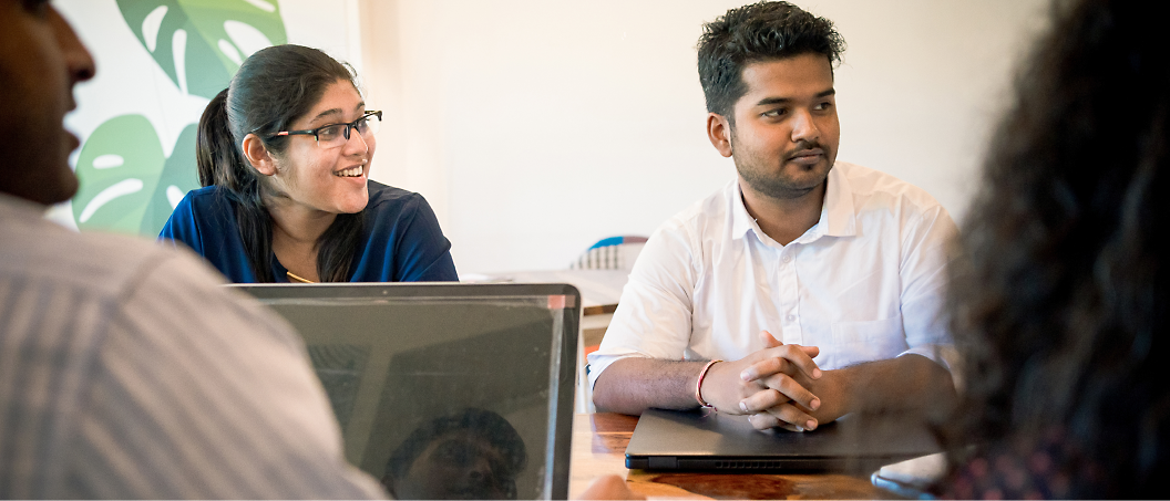 Two individuals are seated at a table during a meeting. The woman on the left is smiling, while the man on the right looks attentive.