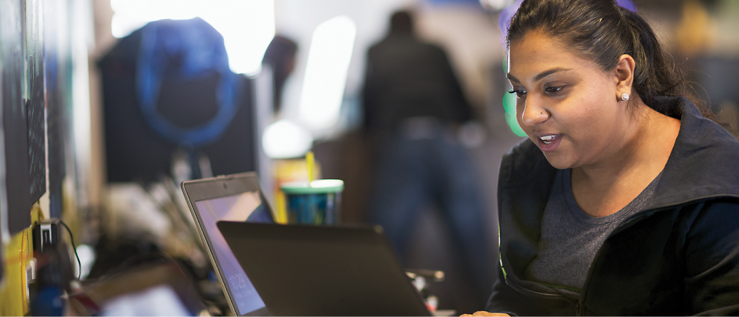 A woman sits at a desk working on a laptop, with another laptop and a drink beside her. She appears focused on her work.