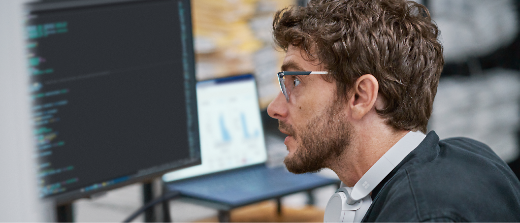 A man wearing glasses and headphones works on a computer with lines of code visible on the screen in front of him.