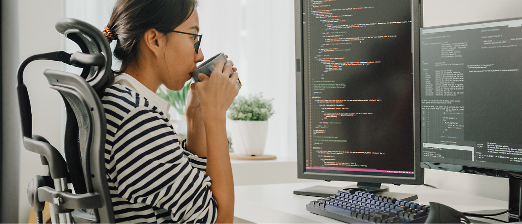 A person sits at a desk, drinking from a cup while looking at two computer monitors displaying code.