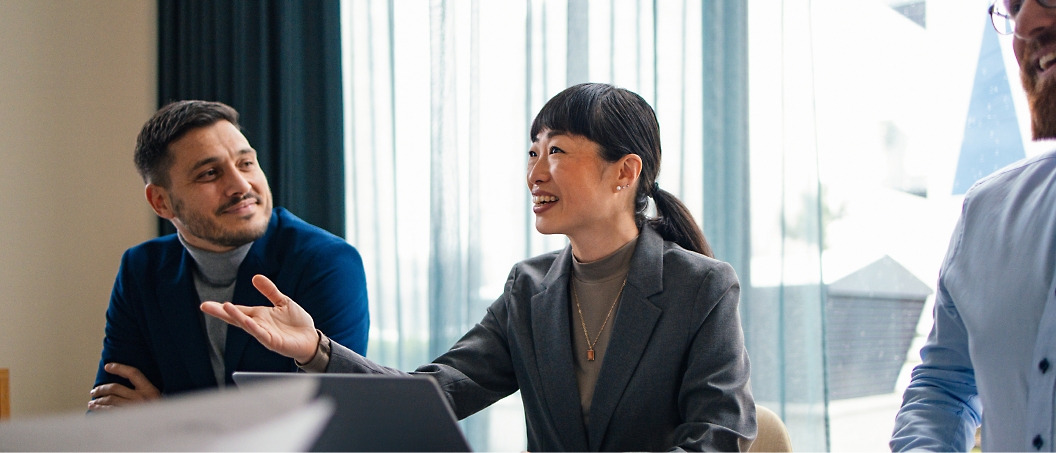 Three people in business attire sit at a table during a meeting. One person is speaking and gesturing with a hand