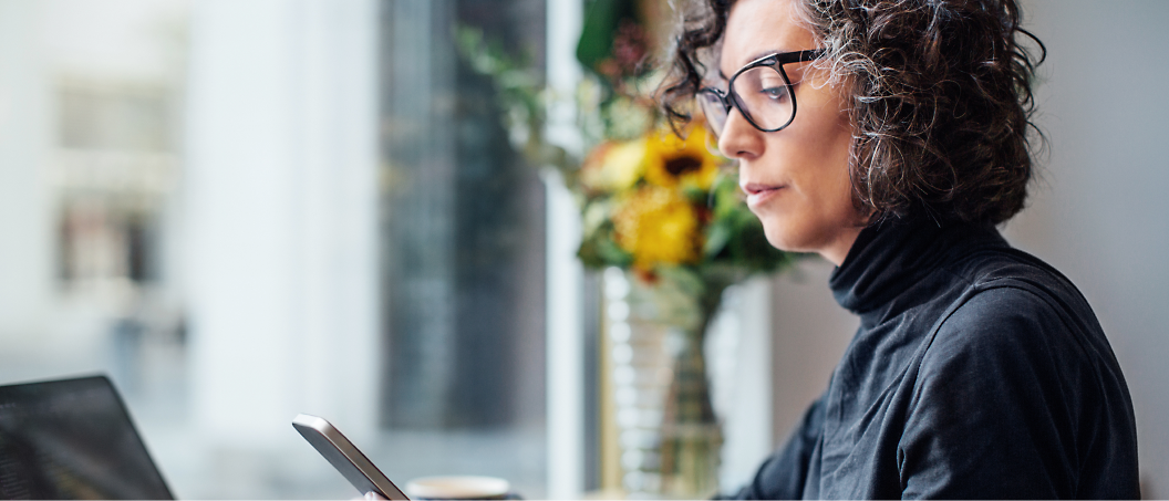 A person with curly hair and glasses sits by a window, looking at a smartphone. 