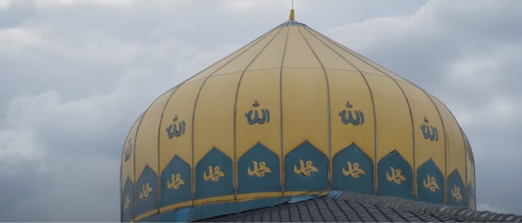 The image shows the dome of a mosque adorned with blue Arabic calligraphy against a cloudy sky.