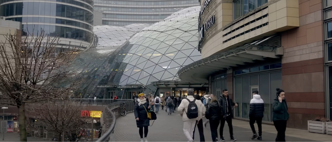 People walk outside a modern building with a glass dome roof. Bare trees and a bicycle are in the foreground.