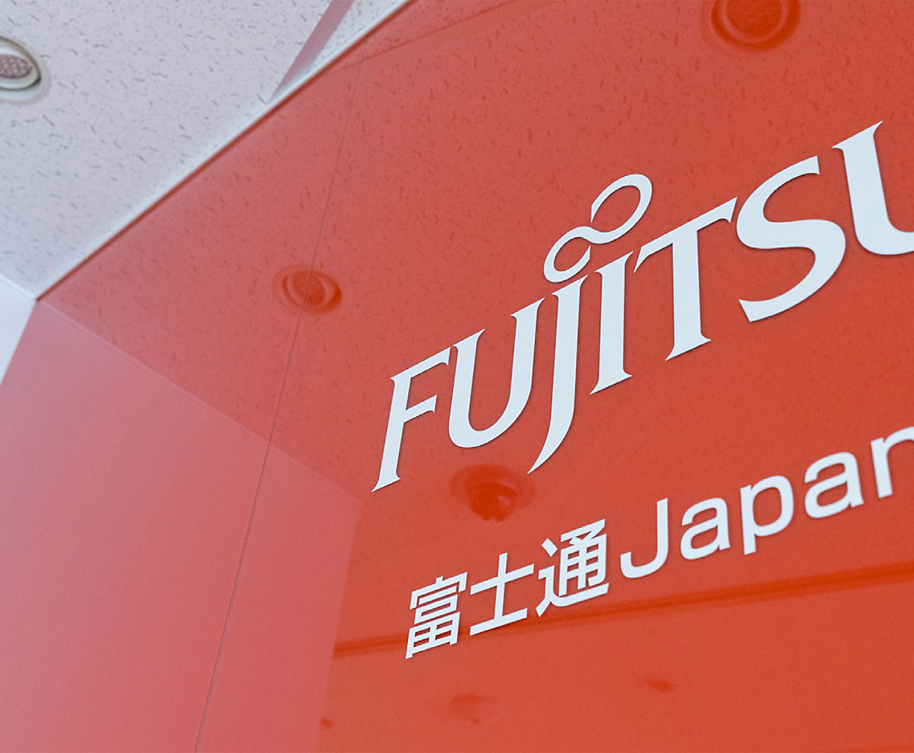 Delvis närbild av en Fujitsu-skylt med logotyp och japansk text på en röd bakgrund, vinklat perspektiv 