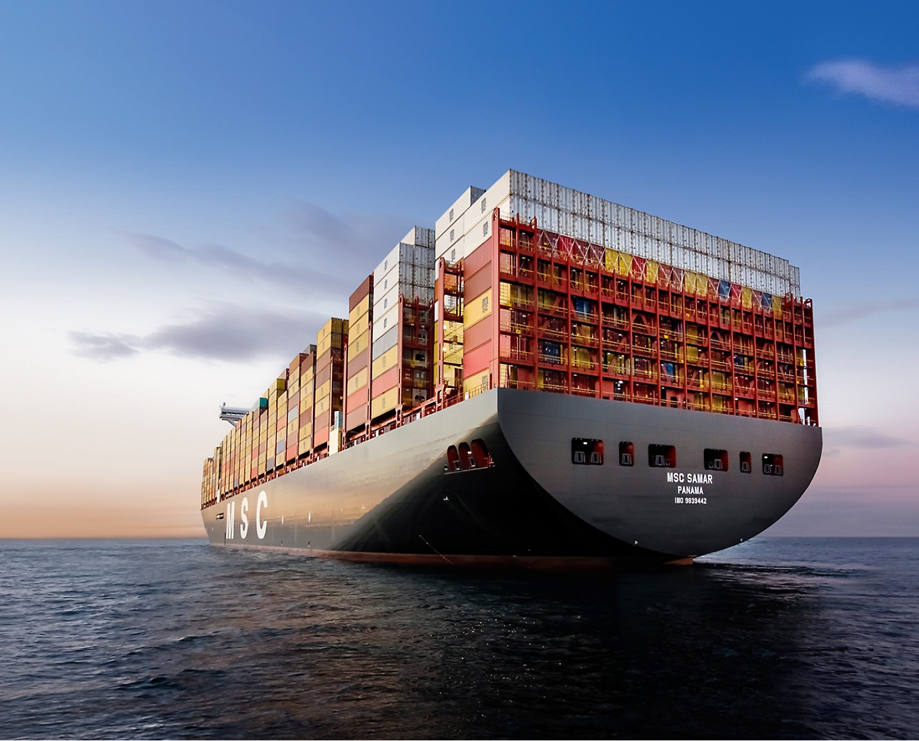 A large cargo ship loaded with multicolored shipping containers sails on calm waters under a clear sky.