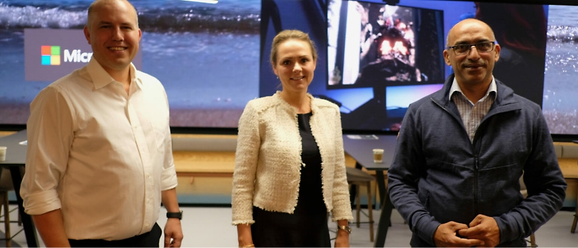 Three people are standing and smiling in front of a large screen displaying various images, including a Microsoft logo. 