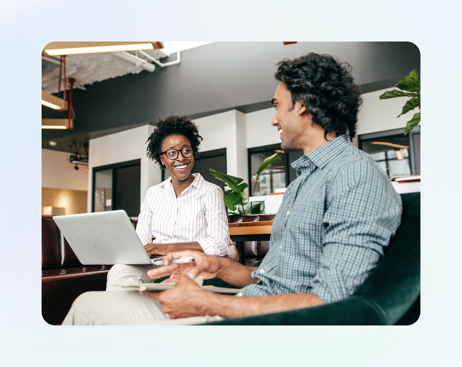 Two people are sitting in a modern office space, one holding a laptop and the other using a tablet, engaged in a conversation.