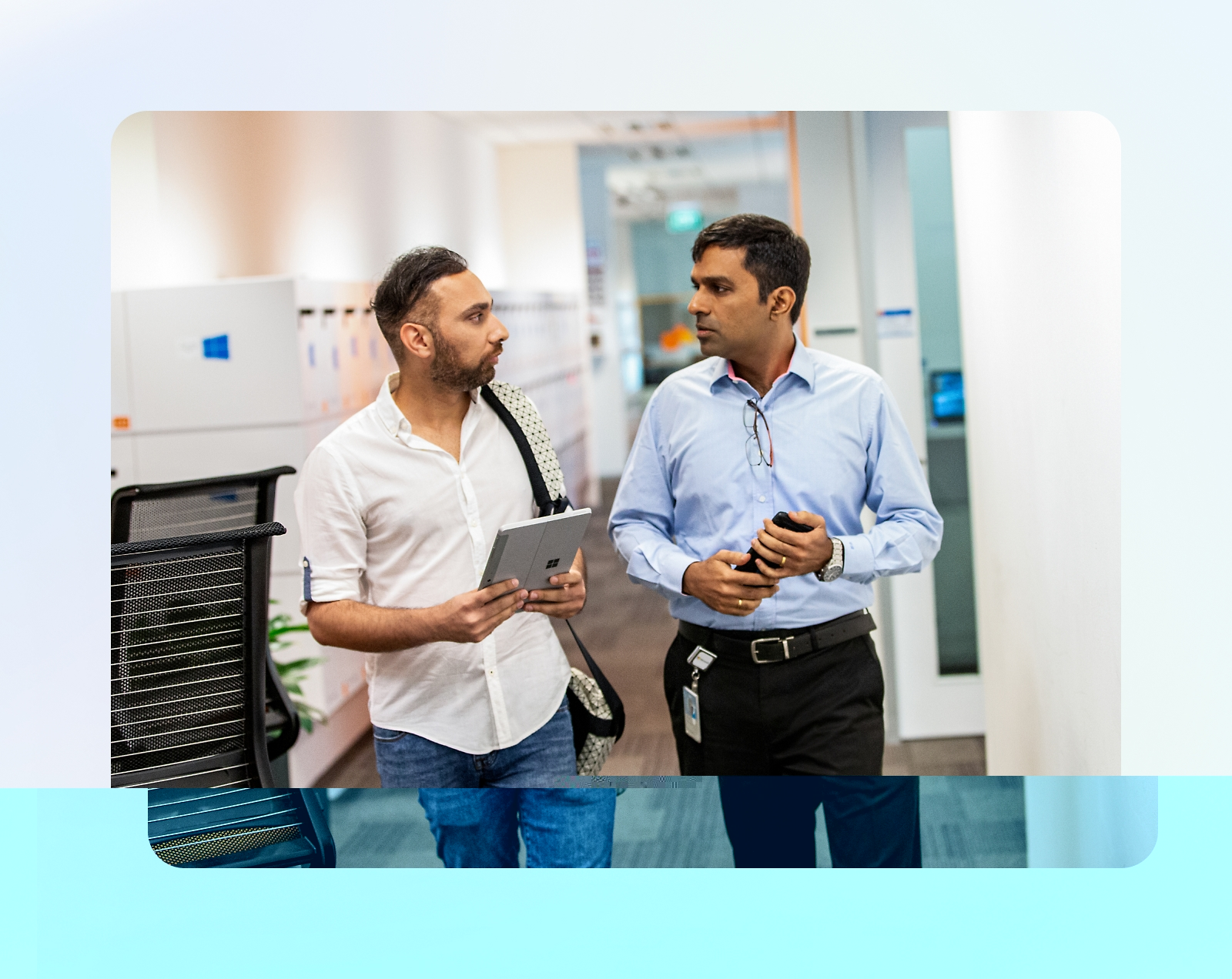 Two men walking down an office corridor, engaged in conversation. One carries a tablet and the other holds a coffee cup.