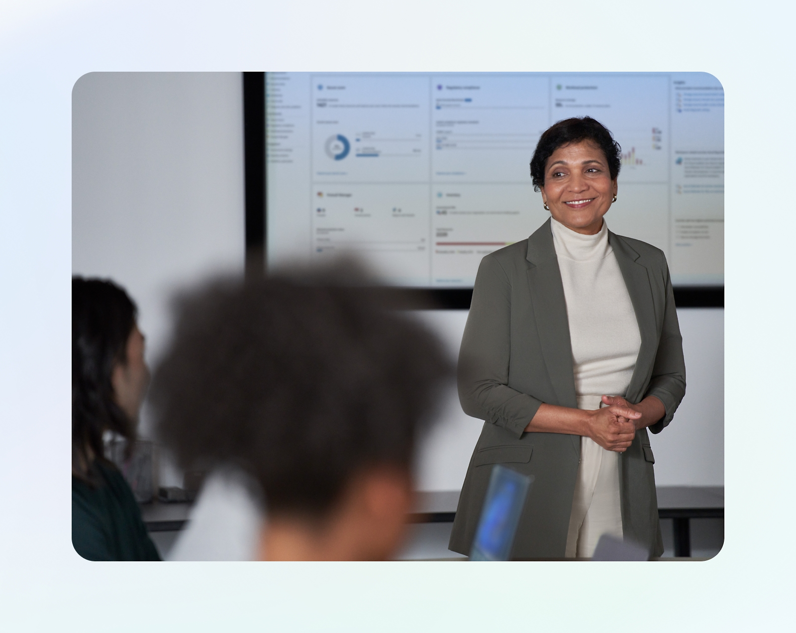 A person in business attire stands in front of a presentation screen, smiling, while two blurred individuals sit facing them.