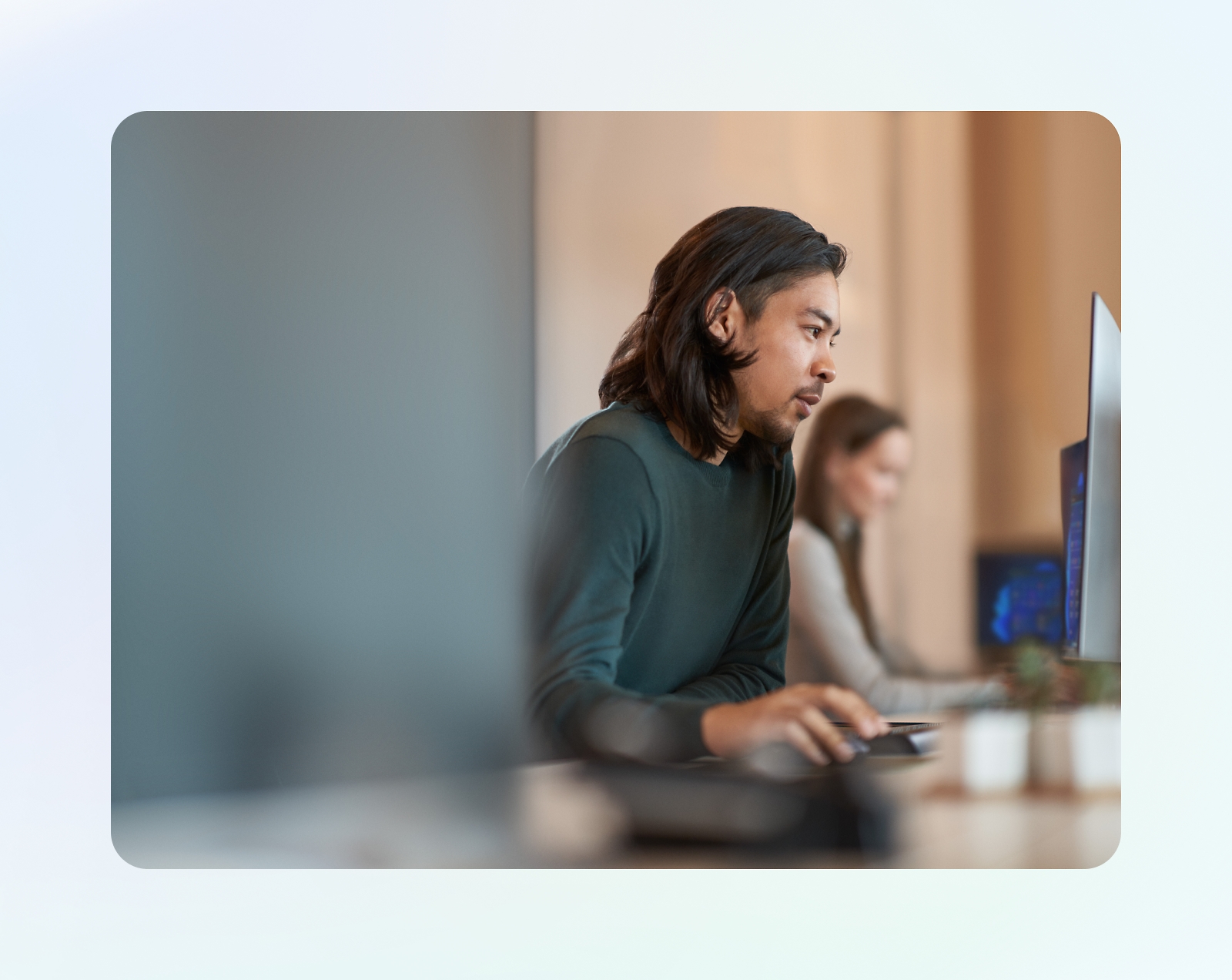 A person with long hair is working on a computer in an office. Another person is working at a computer in the background.