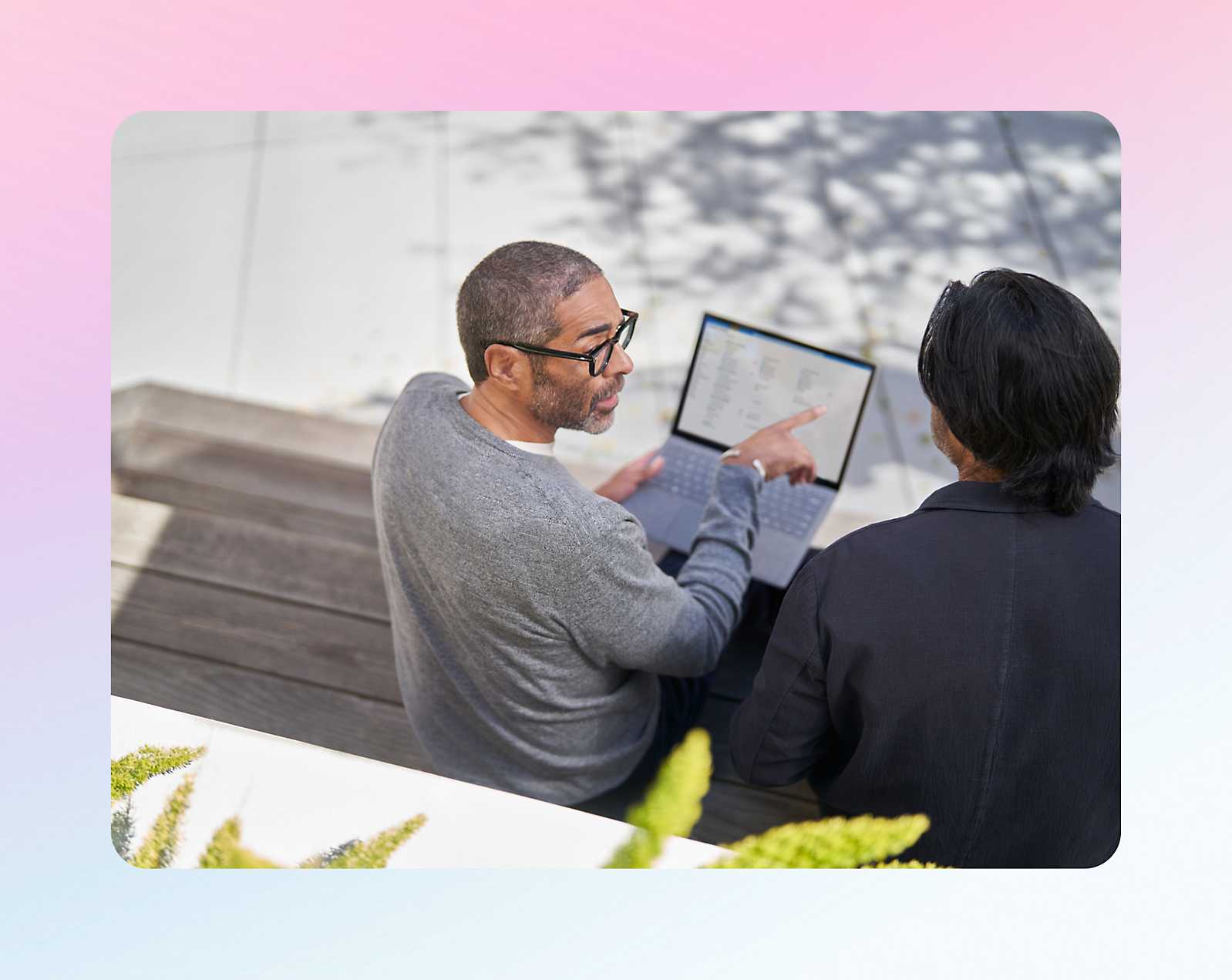 Two people sitting outdoors, one pointing at a laptop screen. The background is blurred with soft pastel hues.