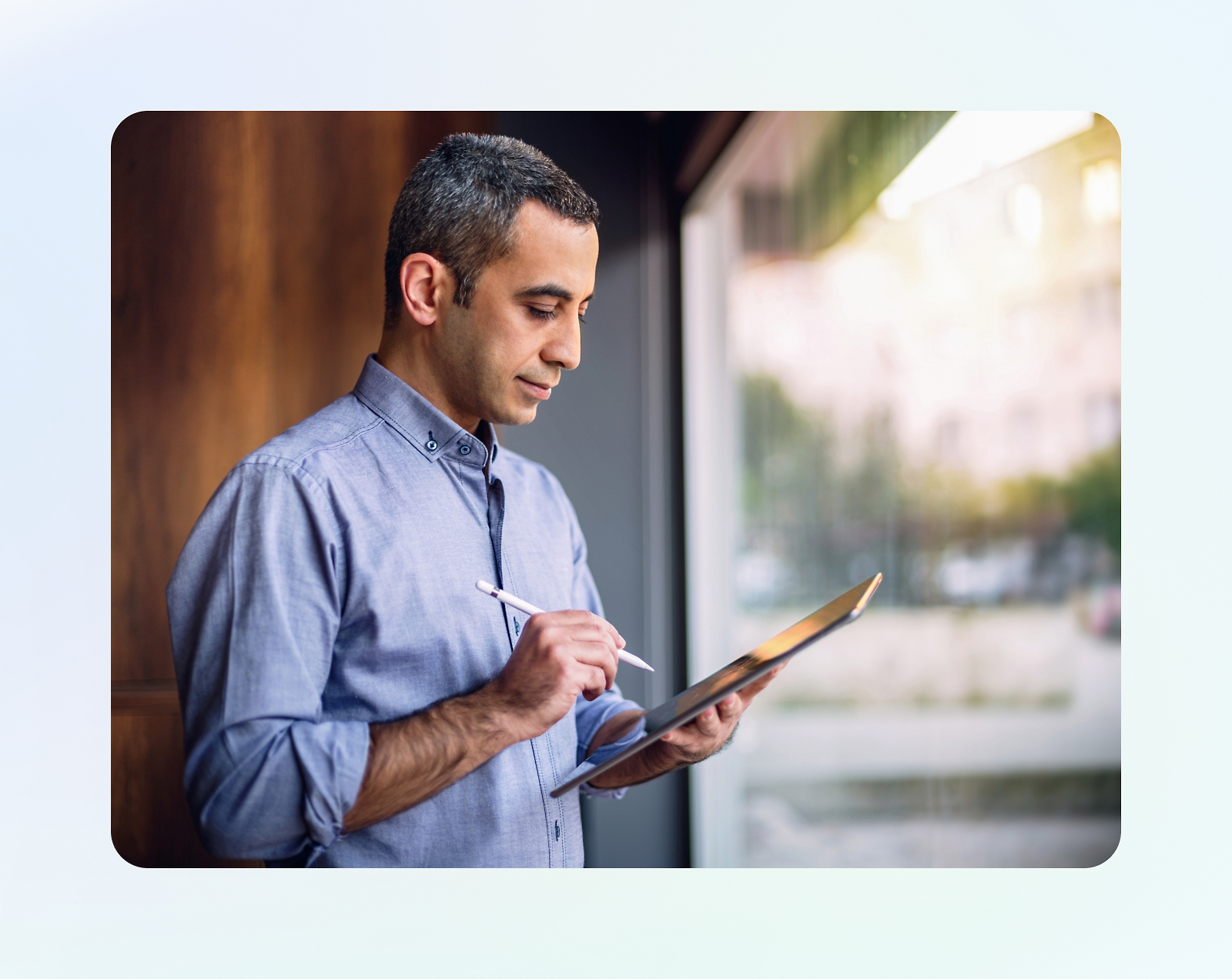 Eine Person in einem blauen Hemd steht in der Nähe eines Fensters und hält ein Tablet mit einem Stift. Der Hintergrund enthält eine verschwommene Szene.