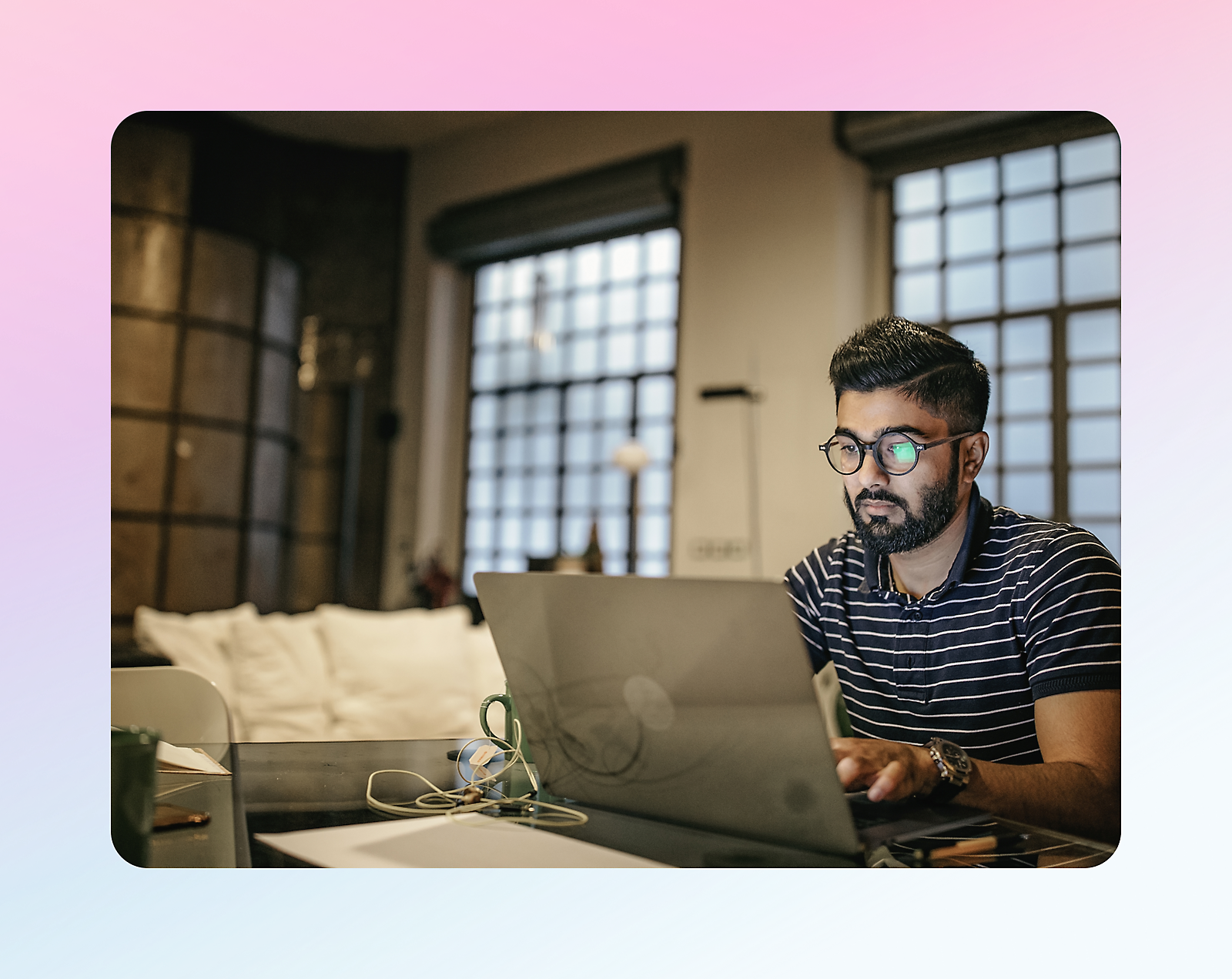 A man wearing glasses and a striped shirt works on a laptop at a desk in a modern room with large windows.