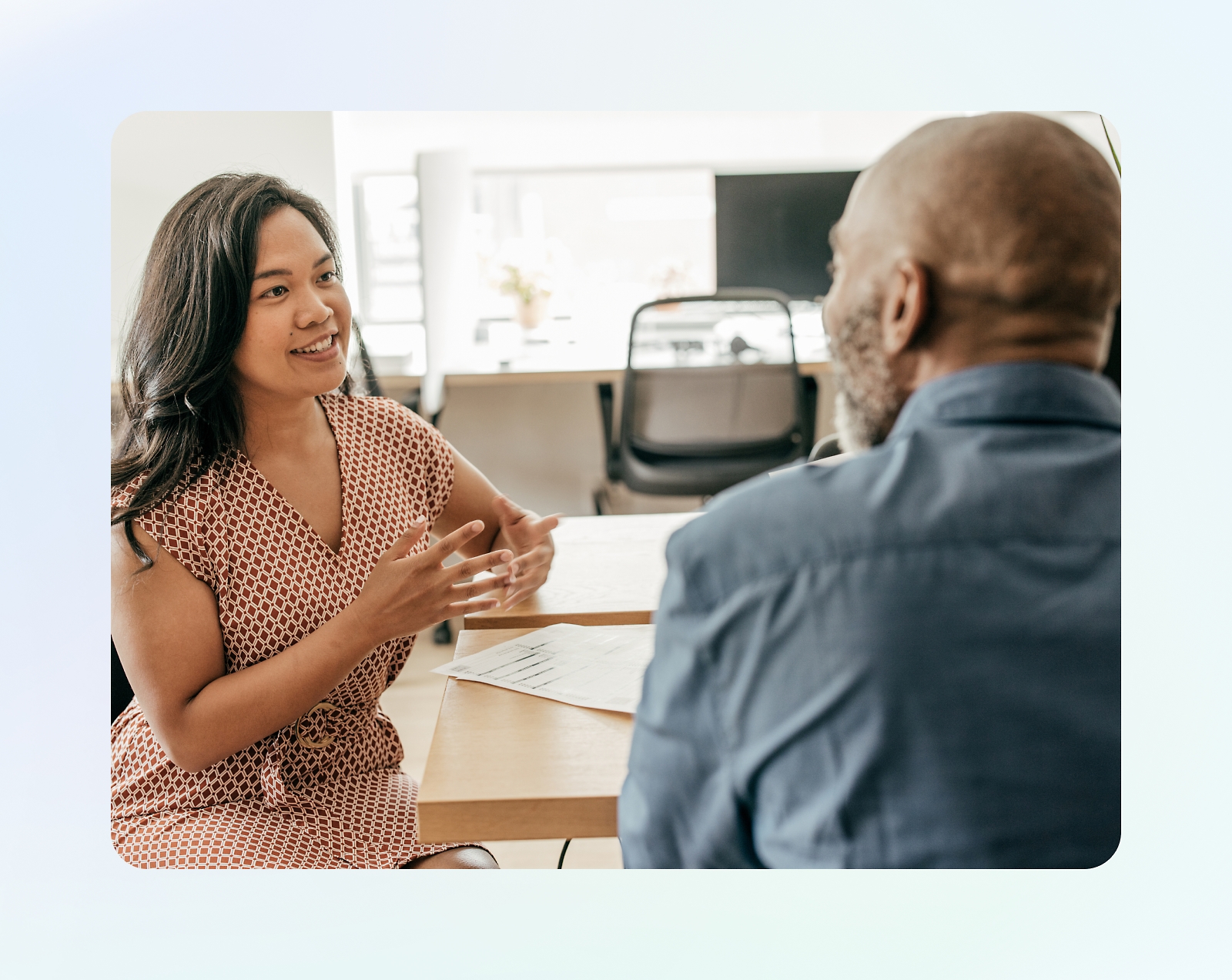 A woman and a man are sitting at a table, engaged in a conversation. The woman is gesturing with her hands 