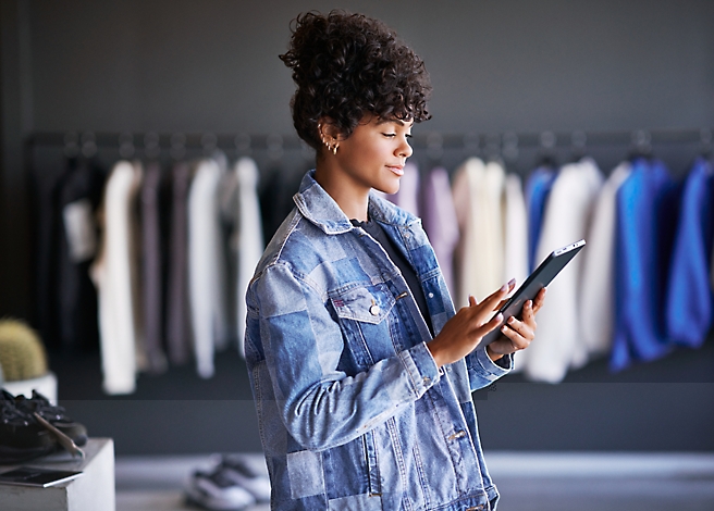 A person with curly hair is standing in a clothing store, wearing a denim jacket and using a tablet