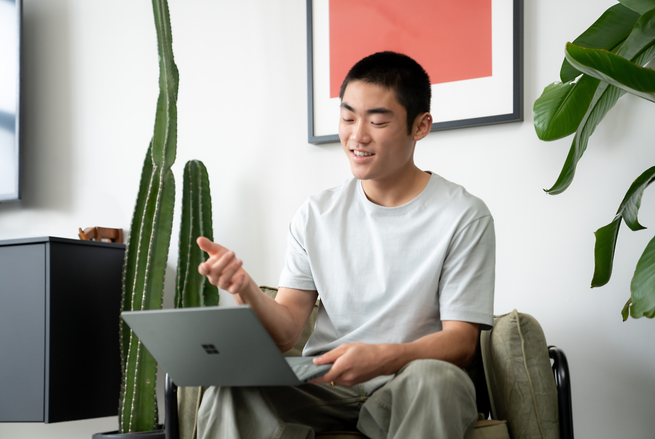 A person is sitting in a chair holding a laptop and gesturing with their hand.