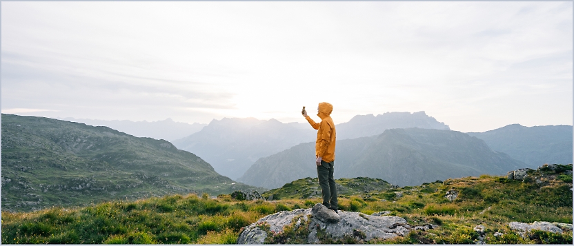 A person stands on a rock in a mountainous landscape, appearing to take a photo or video with a smartphone.